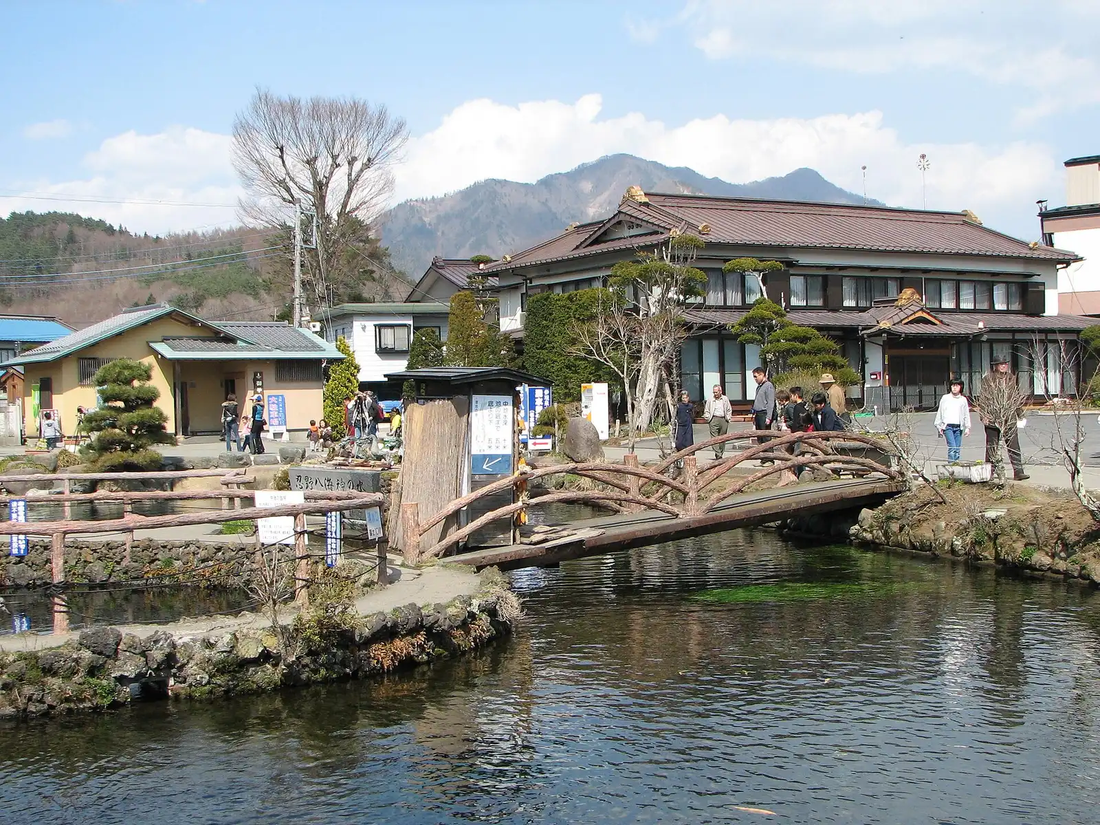 Rustic wooden bridge over a spring-fed pond at Oshino Hakkai, old inn and hills behind